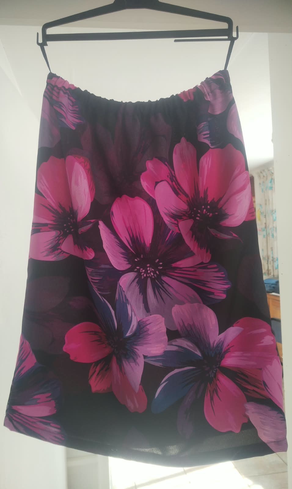 A knee-length skirt in dark fabric with large pink and purple flowers, with a gathered elasticated waist, hanging on a hanger.