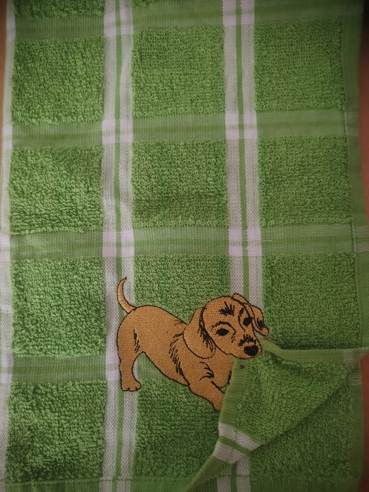 A tan cartoon sausage dog with big puppy eyes embroidered on a green and white checked towel.