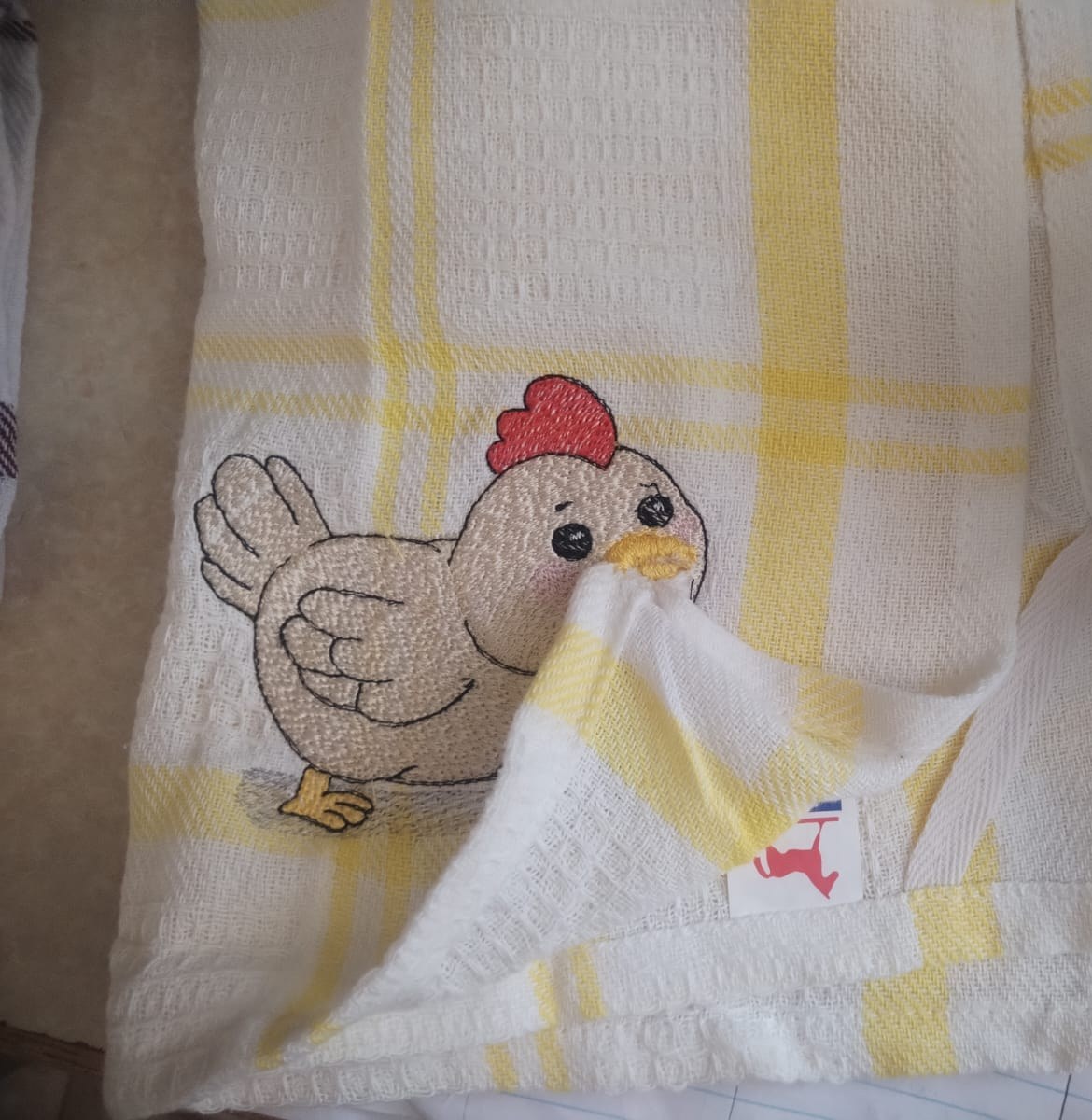 A chubby cartoon hen with a red comb embroidered on a cream and yellow checked tea towel.